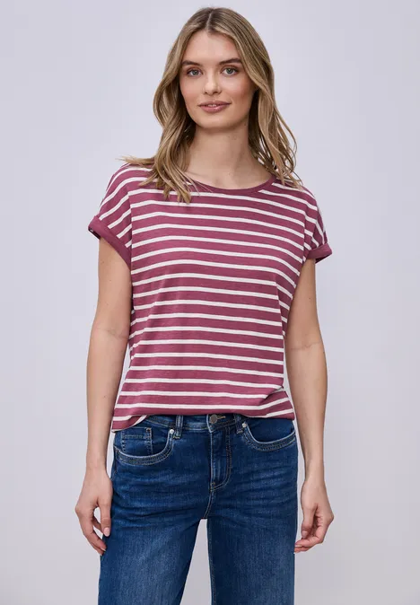 Frau steht vor hellem Hintergrund in gestreiftem rosa-weißem T-Shirt und blauen Jeans.